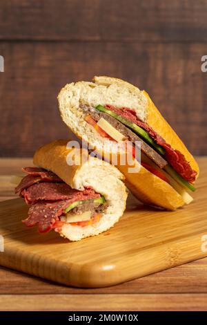 Baguette-Sandwich. Sandwich mit Salami, Roastbeef, Cheddar, Tomaten und Gurke auf Holzhintergrund Stockfoto