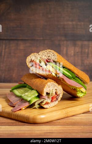 Baguette-Sandwich. Sandwich mit Salami, geräuchertem Fleisch, Oliven, Salat, Tomaten und Gurke auf Holzhintergrund Stockfoto