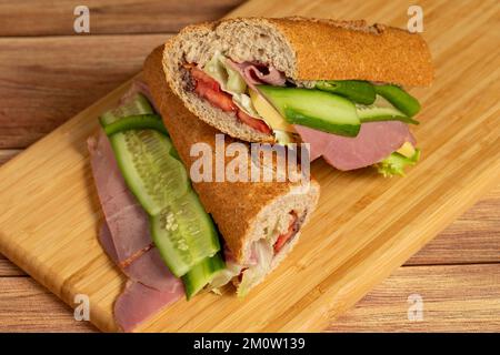 Baguette-Sandwich. Sandwich mit Salami, geräuchertem Fleisch, Oliven, Salat, Tomaten und Gurke auf Holzhintergrund Stockfoto