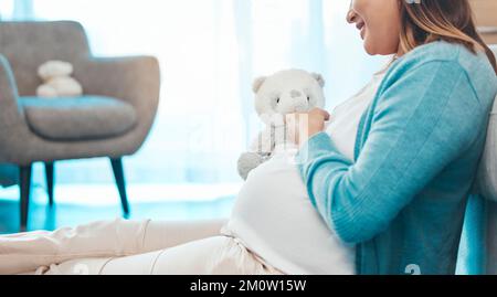 Entspann dich, fröhliche und schwangere Frau mit Teddybär in der Kinderkrippe, die sich auf die Mutterschaft freut. Schwangerschaftsbauch eines Mädchens, das sich zu Hause auf dem Boden ruht, mit einem Lächeln Stockfoto