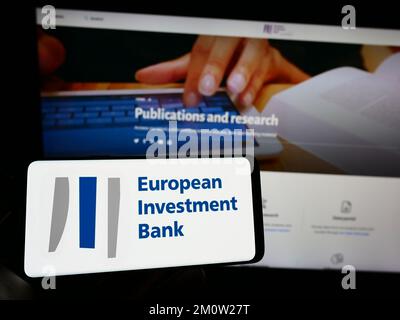 Person, die ein Mobiltelefon mit dem Logo der EU-Institution Europäische Investitionsbank (EIB) auf dem Bildschirm vor der Webseite hält. Konzentrieren Sie sich auf das Display des Telefons. Stockfoto