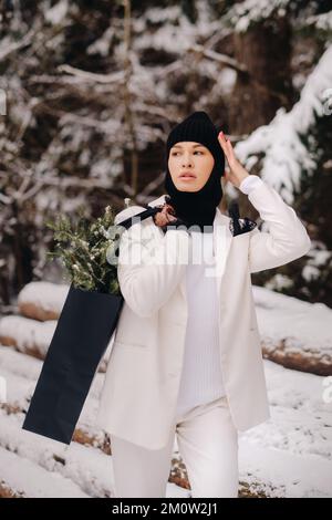 Ein Mädchen in einem weißen Anzug und Balaklava mit einem Paket Weihnachtsbäume im Winterwald an Silvester. Neujahrskonzept Stockfoto