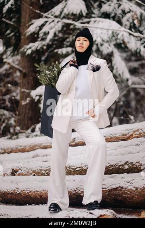 Ein Mädchen in einem weißen Anzug und Balaklava mit einem Paket Weihnachtsbäume im Winterwald an Silvester. Neujahrskonzept Stockfoto