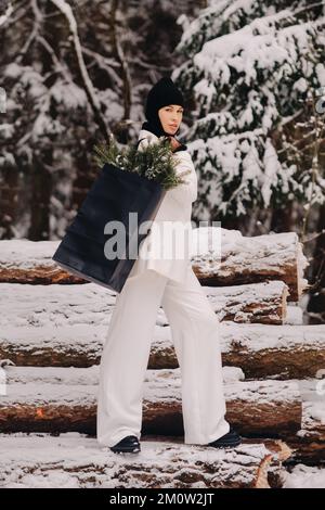 Ein Mädchen in einem weißen Anzug und Balaklava mit einem Paket Weihnachtsbäume im Winterwald an Silvester. Neujahrskonzept Stockfoto