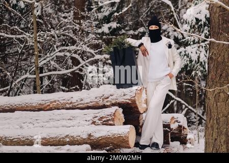 Ein Mädchen in einem weißen Anzug und Balaklava mit einem Paket Weihnachtsbäume im Winterwald an Silvester. Neujahrskonzept Stockfoto