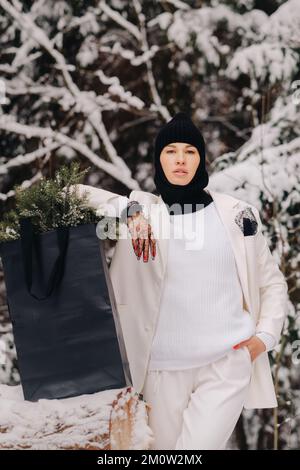 Ein Mädchen in einem weißen Anzug und Balaklava mit einem Paket Weihnachtsbäume im Winterwald an Silvester. Neujahrskonzept Stockfoto