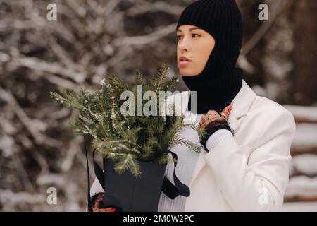 Ein Mädchen in einem weißen Anzug und Balaklava mit einem Paket Weihnachtsbäume im Winterwald an Silvester. Neujahrskonzept Stockfoto