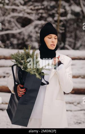 Ein Mädchen in einem weißen Anzug und Balaklava mit einem Paket Weihnachtsbäume im Winterwald an Silvester. Neujahrskonzept Stockfoto