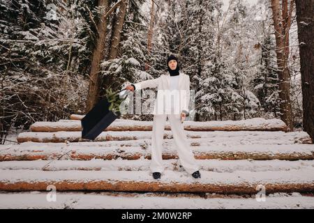 Ein Mädchen in einem weißen Anzug und Balaklava mit einem Paket Weihnachtsbäume im Winterwald an Silvester. Neujahrskonzept Stockfoto