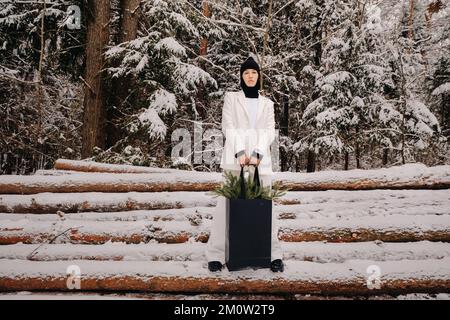 Ein Mädchen in einem weißen Anzug und Balaklava mit einem Paket Weihnachtsbäume im Winterwald an Silvester. Neujahrskonzept Stockfoto