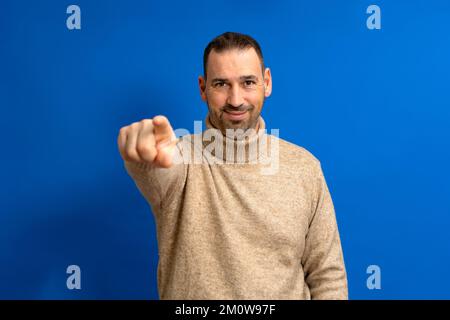 Porträt eines lächelnden hispanischen Mannes in braunem Rollkragenpulli, der mit dem Finger auf die Kamera zeigt, isoliert auf blauem Hintergrund. Stockfoto