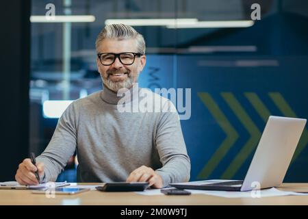 Porträt eines erfolgreichen Geschäftsmanns, eines grauhaarigen Mannes mit Brille, der lächelt und in die Kamera schaut, eines reifen Investorenbosses, der in einem modernen Bürogebäude mit einem Laptop arbeitet. Stockfoto