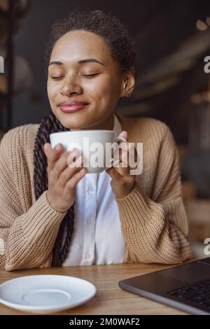 Aus nächster Nähe eine hübsche ethnische Frau, die einen Becher mit heißem Getränk hält und das Aroma von Cappuccino genießt, während sie im Café chillt Stockfoto