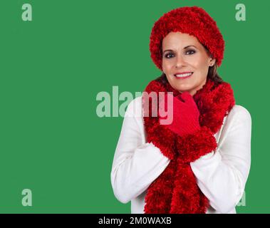 Wunderschöne, reife Frau trägt einen weißen Pullover mit rotem Hut, Schal und Handschuhen vor grünem Hintergrund für die Weihnachtszeit Stockfoto