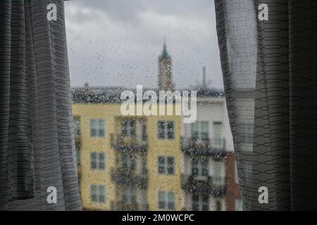 Vorhänge umgeben einen Blick auf die Stadt aus einem Fenster, das an einem bewölkten Tag von Regentropfen bedeckt ist. Stockfoto