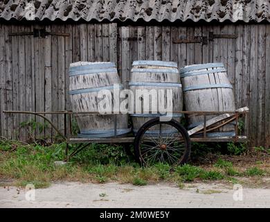 Drei alte Fässer auf einem Holzwagen an der Holzwand eines Schuppens während des Tages Stockfoto