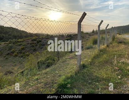 Eisenzaun und Stacheldraht parallel zur Schotterstraße. Sonnenuntergang auf dem Land. Selektiver Fokus auf Zaunwand. Stockfoto