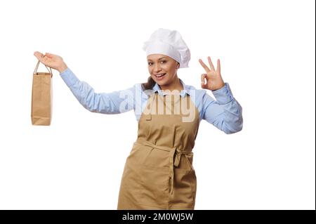 Hübsche Kellnerin mit Koch-Hut und beiger Schürze, hält Papiertüte mit Essen zum Mitnehmen, zeigt OK-Schild, weißer Hintergrund Stockfoto