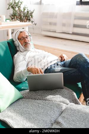 Porträt alter alter alter alter Mann mit Kopfhörern, der zu Hause Musik auf einem Laptop hört - Konzept für aktive Senioren Stockfoto