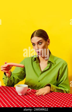 Emotionale junge Frau Vintage Kleidung isst Würstchen mit Kaffee, Cappuccino auf roter Tischdecke auf gelbem Hintergrund. Pop-Art-Fotografie mit Speisen. Stockfoto