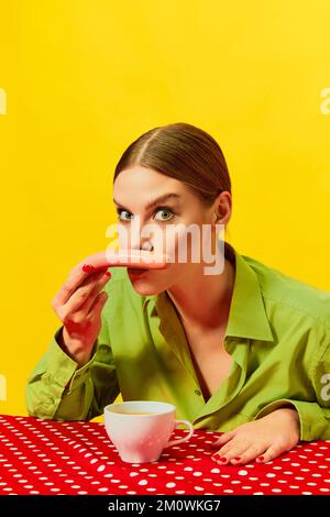 Junge Frau, Vintage-Kleidung, die Würstchen mit Kaffee schnüffelt, Cappuccino auf roter Tischdecke auf gelbem Hintergrund. Pop-Art-Fotografie mit Speisen. Stockfoto