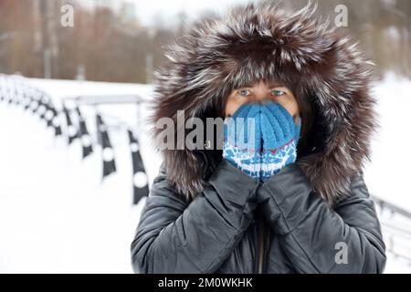 Frostwetter im Winter, Frau im Ledermantel mit Pelzhaube, die während des Schnees auf einer Straße unterwegs ist und ihr Gesicht mit gestrickten Handschuhen mit den Händen bedeckt Stockfoto