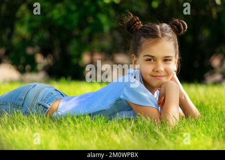 die junge braune weiße Frau liegt auf dem Gras im Park und lächelt mit erhobenem Kopf und Händen. Hochwertiges Foto Stockfoto