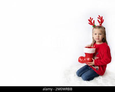 Weihnachten. Ein Mädchen mit Hirschhörnern in einem roten Pullover hält weihnachtsmannstiefel. Banner Stockfoto