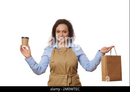 Süße Kellnerin in beigefarbener Küchenschürze, hält eine Tasse Getränke zum Mitnehmen aus Pappe und eine Papiertüte mit frischem Essen Stockfoto