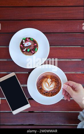 Hand hält eine Tasse heißen Cappuccino-Kaffee zusammen mit einem Teller mit Weihnachtsschokolade und Smartphone Stockfoto