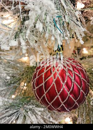 Nahaufnahme eines roten Weihnachtsschmucks mit einem goldenen glitzernden Gittermuster, das an einem weißen Weihnachtsbaum hängt Stockfoto