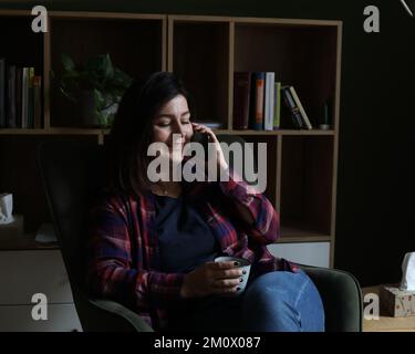 Konzentrierte, lächelnde, ruhige, brünette Geschäftsfrau in einem karierten Hemd, die Tee oder Kaffee trinkt, im Büro ein Mobiltelefon spricht Stockfoto