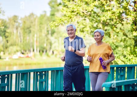 Asiatische und kaukasische Ehefrau und Ehemann mittleren Alters tragen aktive Kleidung, spazieren entlang der Brücke im Sommerpark, genießen Sie morgens einen sportlichen Spaziergang draußen. Stockfoto