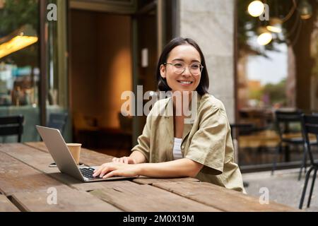 Porträt eines asiatischen Mädchens, das im Café arbeitet, einen Laptop benutzt und draußen auf der Straße sitzt. Digitales Nomaden- und Online-Lernkonzept Stockfoto