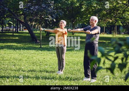 Ältere 60 Ehepartner tragen Sportbekleidung, die sich morgens im Sommerpark mit Gummibändern im Freien üben. Gesunde Lif Stockfoto