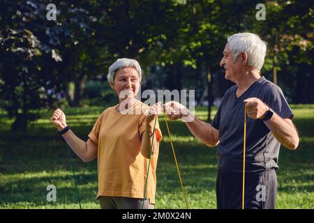 Ältere 60 Ehepartner tragen Sportbekleidung bei Übungen und trainieren morgens im Sommerpark mit Gummibändern im Freien. Gesundes Leben Stockfoto