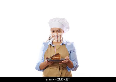 Charmante Hausfrau in beigefarbener Schürze und Hut, Halteteller mit Schokoladenkuchen mit glitzernder Kerze, auf weißem Hintergrund Stockfoto