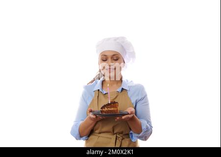 Eine glückliche Frau, die einen Teller mit Schokoladenkuchen hält, Wünsche erfüllt, Kerze ausbläst. Backen für die Feiertage Stockfoto