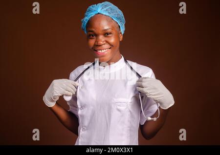 Eine glückliche, junge, schwarze Krankenschwester, die auf eine Kamera auf braunem Hintergrund schaut. Stockfoto