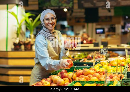 Eine junge, wunderschöne Hidschabi-Muslime, eine Supermarktkraft, legt Früchte in der Abteilung. Er hält Äpfel in den Händen, sieht in die Kamera, lächelt. Stockfoto