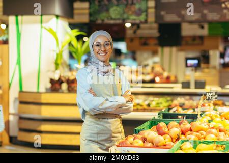 Porträt einer wunderschönen jungen muslimischen Frau in einem Hijab, die in einem Supermarkt arbeitet. Sie steht in der Abteilung für Gemüse und Obst, kreuzt ihre Arme, schaut in die Kamera. Sie lächelt. Stockfoto