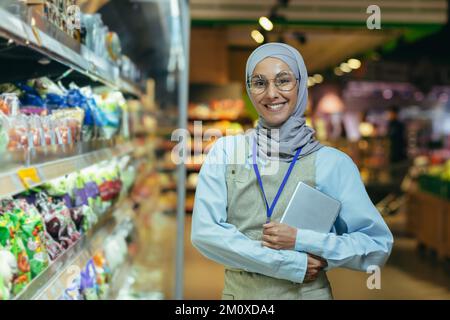 Porträt einer jungen, schönen Frau, einer Supermarktkraft in einem Hijab. Er steht in Uniform mit einem Abzeichen und mit einem Paket in den Händen neben dem Tresen mit Waren, sieht in die Kamera, lächelt. Stockfoto