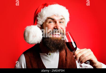 Ein lächelnder bärtiger Mann im Weihnachtsmann trinkt ein Glas Rotwein. Neujahrsparty. Frohe Weihnachten. Stockfoto