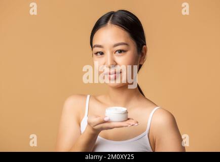 Wunderschöne koreanische Frau hält Feuchtigkeitscreme auf beigefarbenem Hintergrund und schaut auf das Kommen Stockfoto