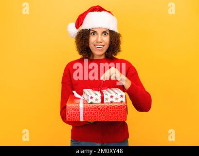 Angenehm aufgeregte glückliche Frau hält ein weihnachtsgeschenk in Händen. Frau mit weihnachtsmann-Hut auf orangefarbenem Hintergrund, lächelnd Stockfoto