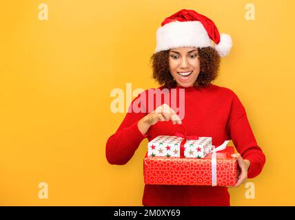 Angenehm aufgeregte glückliche Frau hält ein weihnachtsgeschenk in Händen. Frau mit weihnachtsmann-Hut auf orangefarbenem Hintergrund, lächelnd Stockfoto