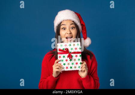 Angenehm aufgeregte glückliche Frau hält ein weihnachtsgeschenk in Händen. Schöne Frau mit weihnachtsmann-Hut auf blauem Hintergrund Stockfoto