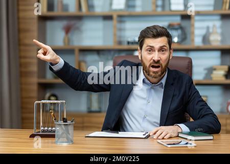 Der wütende Chef ruft vor der Kamera, der Mitarbeiter zeigt mit dem Finger zur Seite und entlässt den Untergebenen, einen Geschäftsmann in einem Geschäftsanzug, der an einem Tisch in einem modernen Büro sitzt. Stockfoto