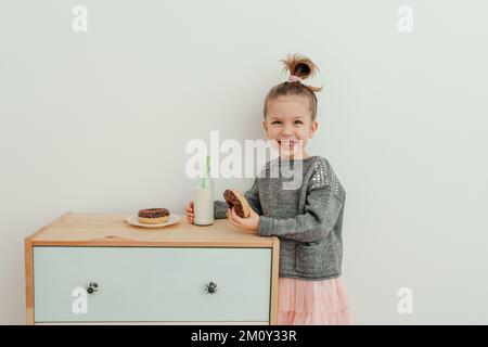 Das hübsche kleine Mädchen isst Schokoladendonut mit einer Flasche Milch. Portrait eines süßen, stilvollen Mädchens, das zufrieden ist, Donuts zu essen. Sweets-Lover-Konzept Stockfoto