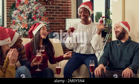 Fröhliche Leute, die ein Glas Wein trinken und sich über die weihnachtsfeier im Büro mit Weihnachtsbaum und Dekor freuen. Verschiedene Kollegen feiern Winterferien und stoßen an. Stockfoto